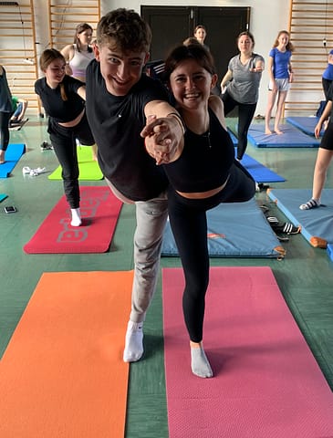 Zwei Jugendliche machen Yoga. Sie machen eine Partnerübung und lachen dabei.